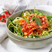 Schüssel gefüllt mit Zucchininudeln, Rucola und Tomatensauce auf weißem Untergrund.