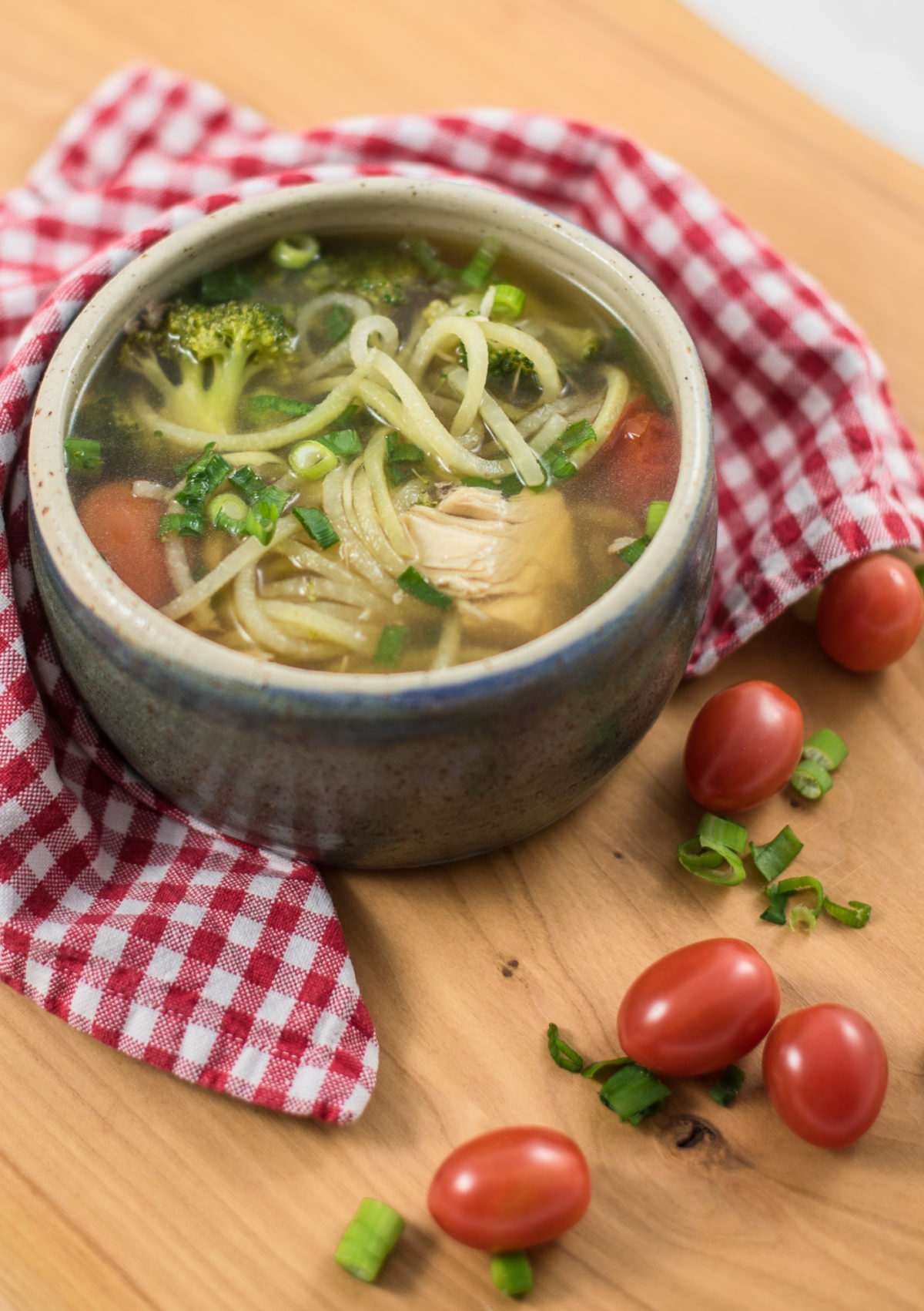 Suppenschüssel mit klarer Brühe und Suppeneinlage auf Holztisch mit rot kariertem Tuch und Tomaten