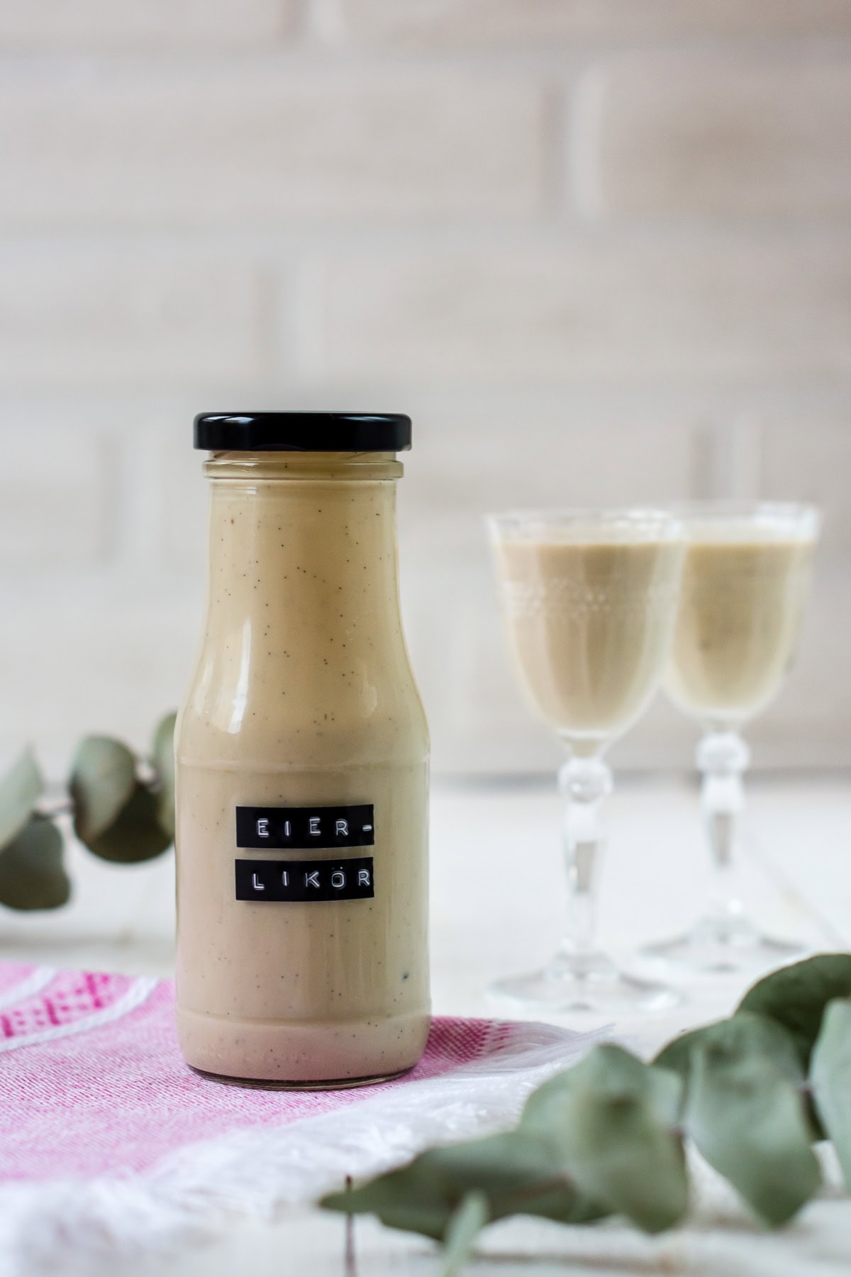 Pinne das Foto, um das Rezept für selbstgemachten Eierlikör nicht aus den Augen zu verlieren! Mit Eierlikör beschriftetes und gefülltes Glas, im Hintergrund zwei Likörgläser mit Eierlikör