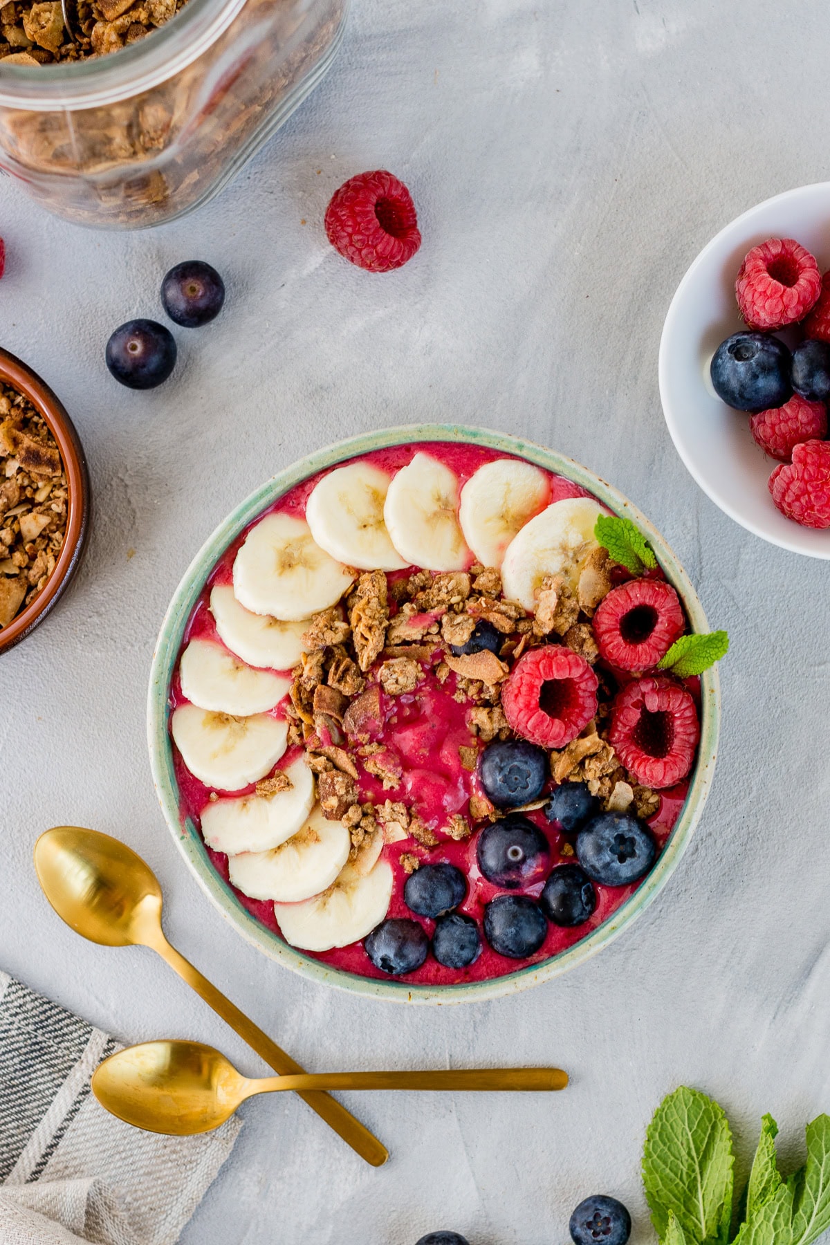 Topshot von einer Schüssel mit pinker Smoothie Bowl getoppt mit Bananenscheiben, Himbeeren, Heidelbeeren und Knuspermüsli.