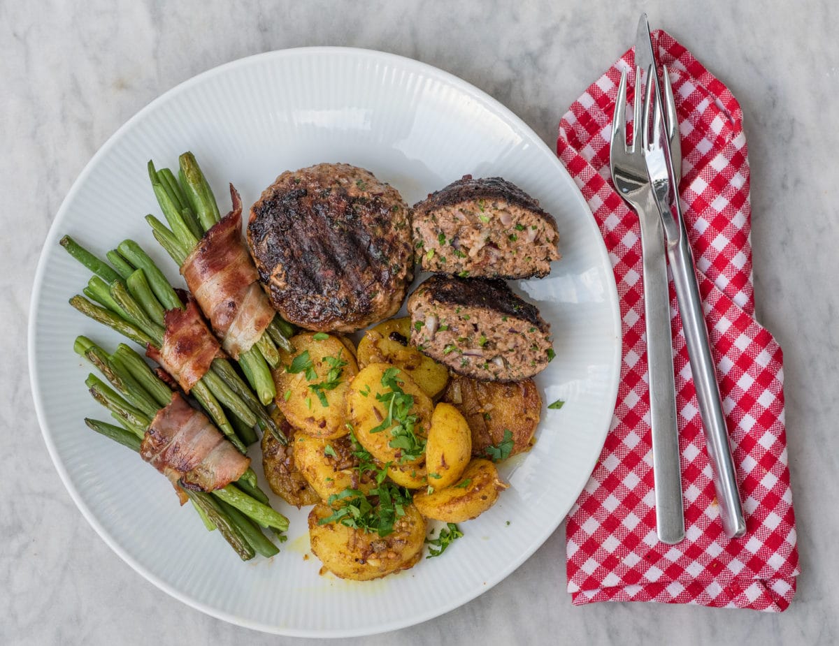 Teller mit Frikadellen, Speckbohnen und Kartoffeln und Besteck rechts davon auf Serviette liegend.