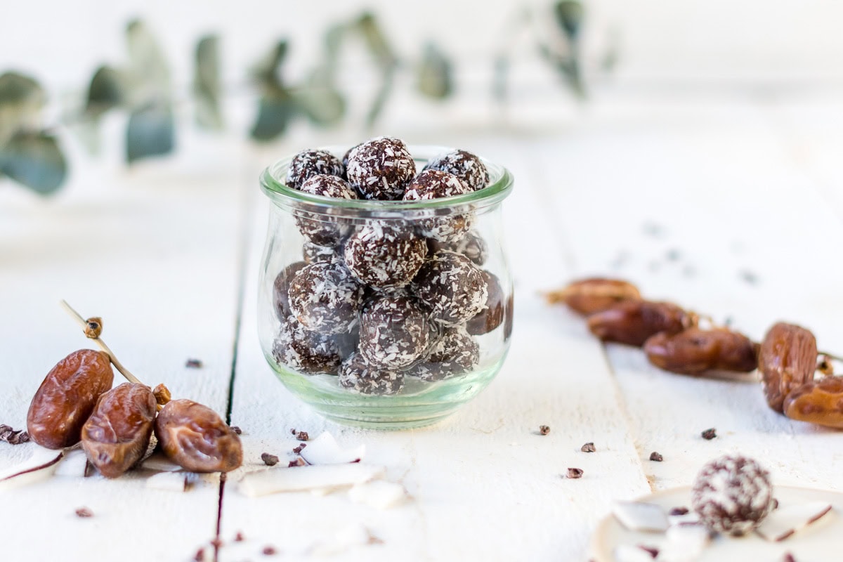 Glas gefüllt mit Schokoladen Bliss Balls und Kokosstreuseln auf weißem Untergrund und Datteln daneben.