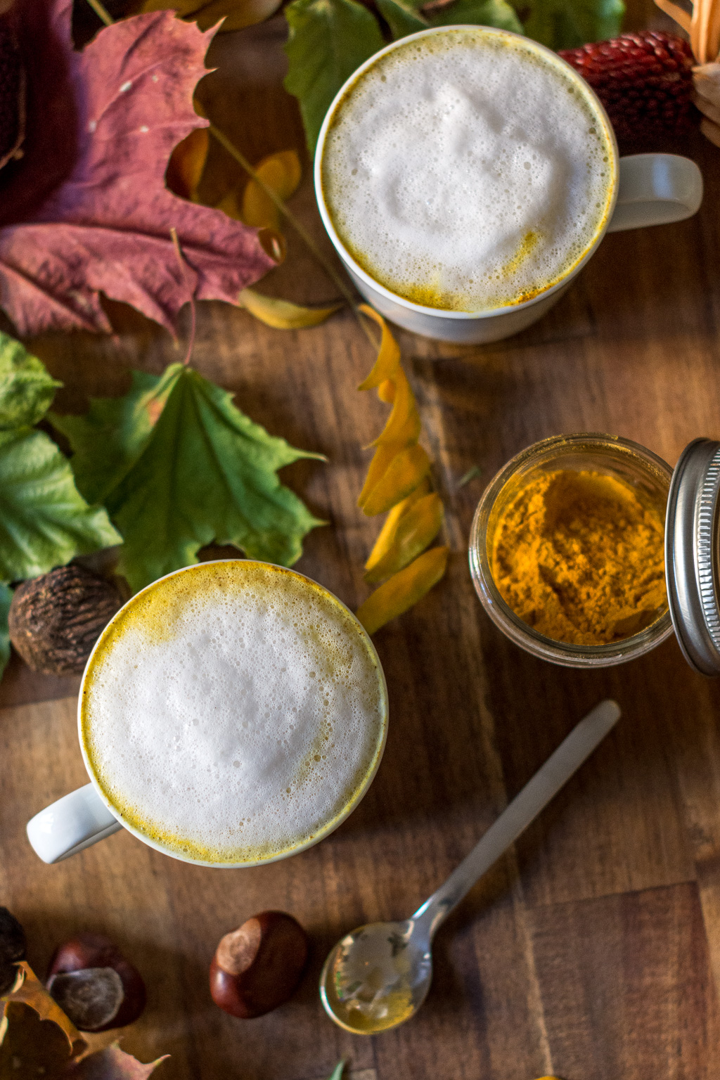 Topshot von zwei Tassen mit Kurkuma Latte gefüllt und mit herbstlichen Blättern dekoriert