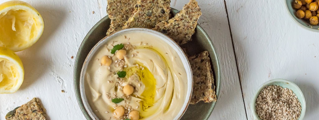 Topshot von Hummus in einer Schale mit Kichererbsen, Knäckebrot, Zitronen und Kernen auf weißem Holzuntergrund dekoriert