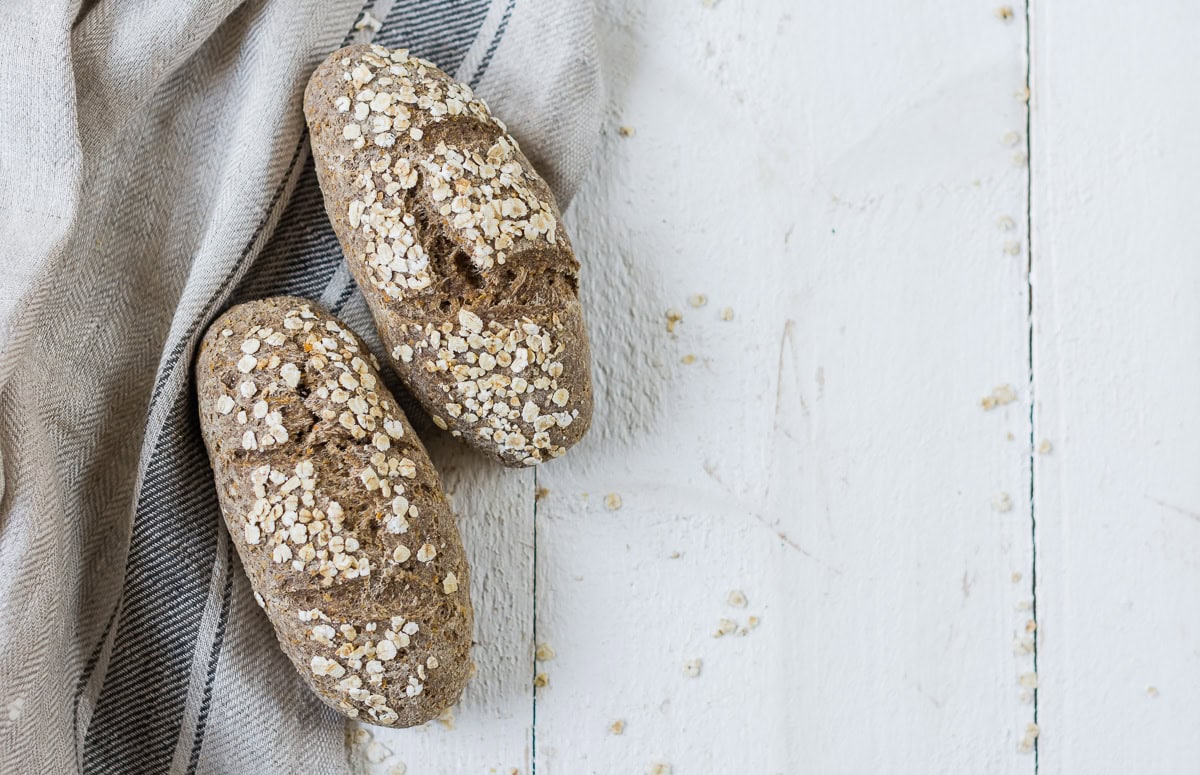 Zwei dunkle Brötchen mit Haferflocken bestreut auf weißem Holzuntergrund