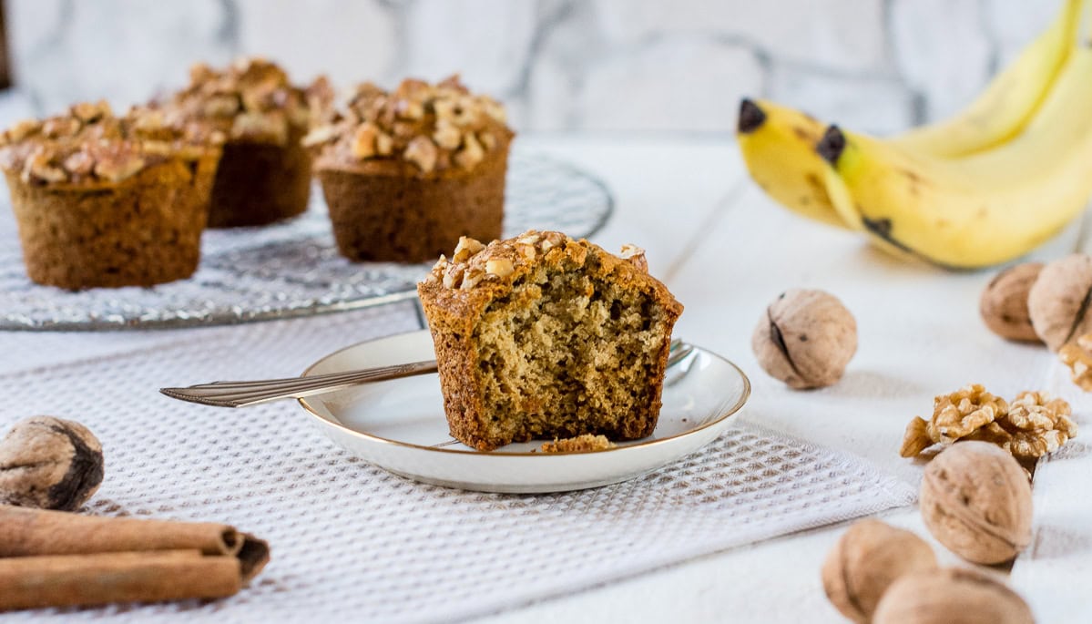 Glutenfreie Bananenmuffins mit Walnüssen und Zimt auf kleinem Teller mit Bananen und Muffins im Hintergrund.