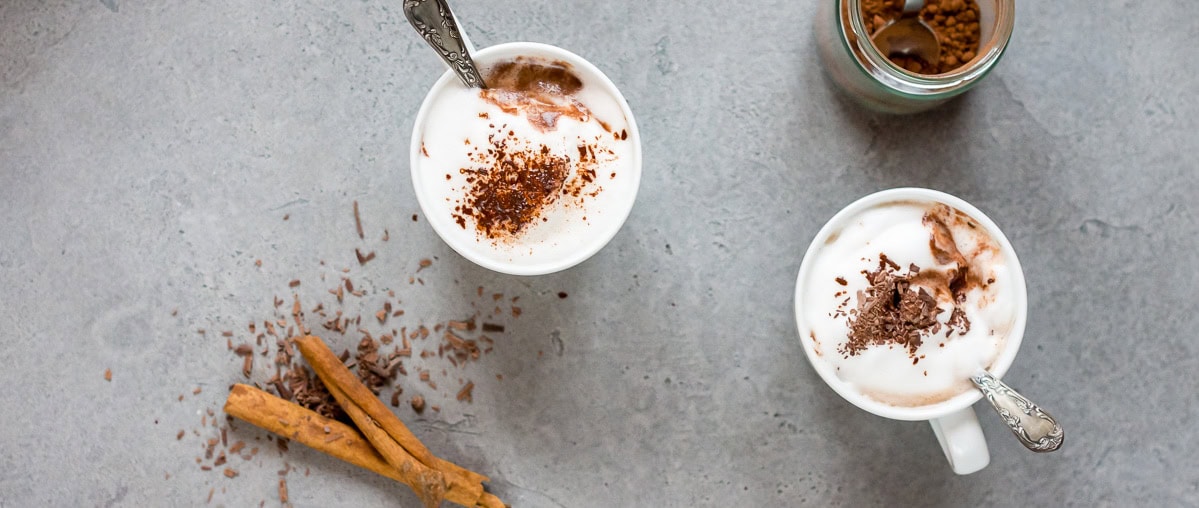 Pinne das Foto, um das Rezept für selbstgemachte, heiße Schokolade zu speichern! Bild von oben von zwei Tassen gefüllt mit heißer Schokolade, Milchschaum und geraspelter Schokolade. Daneben zwei Zimtstangen.