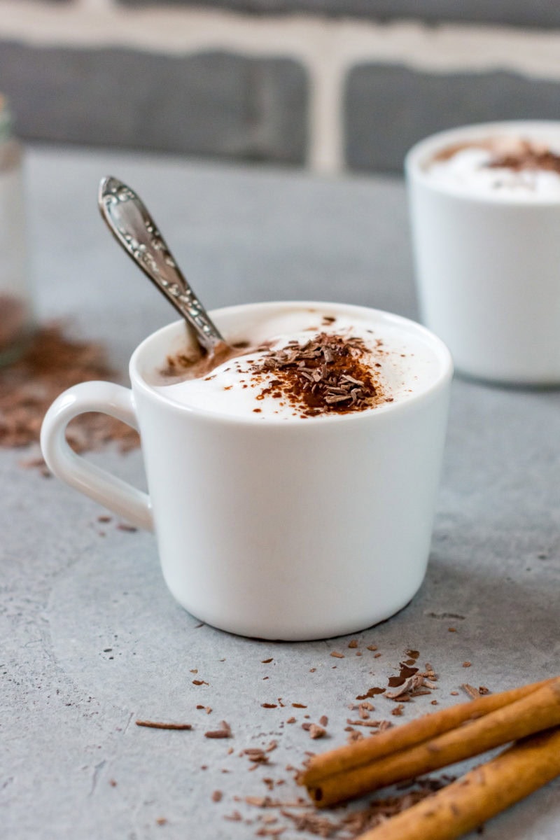 Weiße Tasse mit Milchschaum, einem Teelöffel und geraspelter Schokolade.