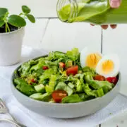 Teller mit grünem Salat, Tomaten und Eiern. Eine Hand hält eine Flasche mit grünem Salatdressing.