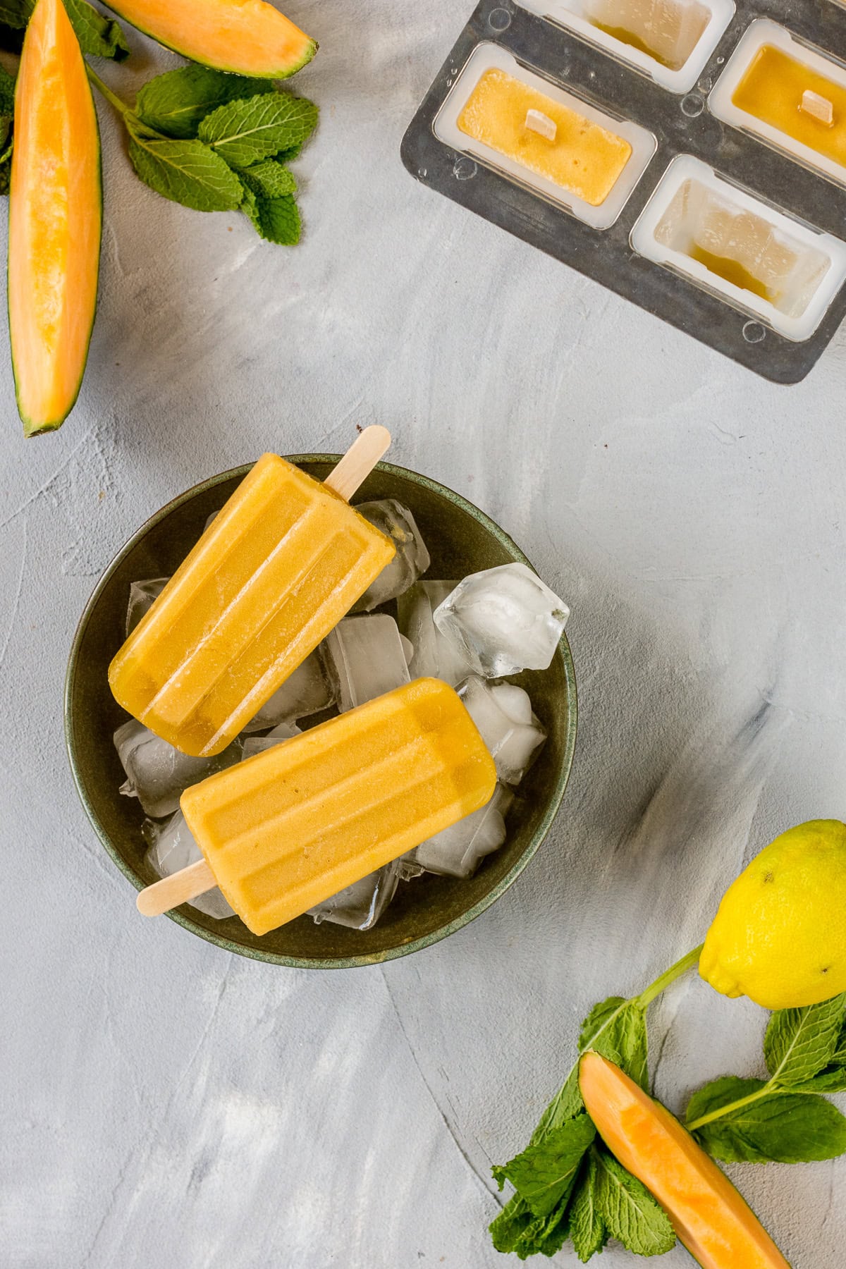 Bild von oben von einer Schüssel mit Eiswürfel in der zwei gelbe Eis am Stiel liegen. Melonenstücke und eine Zitrone liegen daneben.