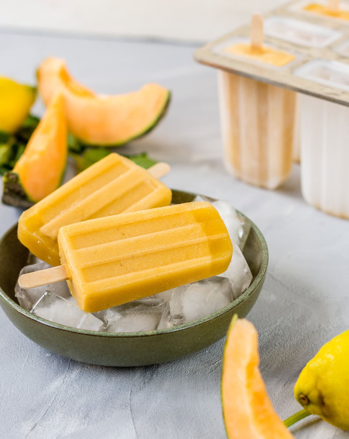 Zwei gelbe Eis am Stiel auf Eiswürfeln in einer Schüssel. Im Hintergrund liegen aufgeschnittene Melonenstücke.