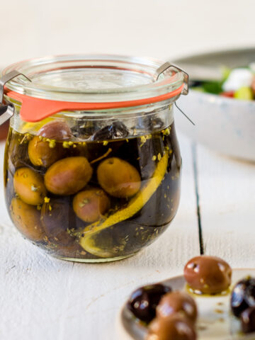 Glas mit eingelegten Oliven und ein Teller mit einem Teelöffel und Oliven auf weißem Untergrund.