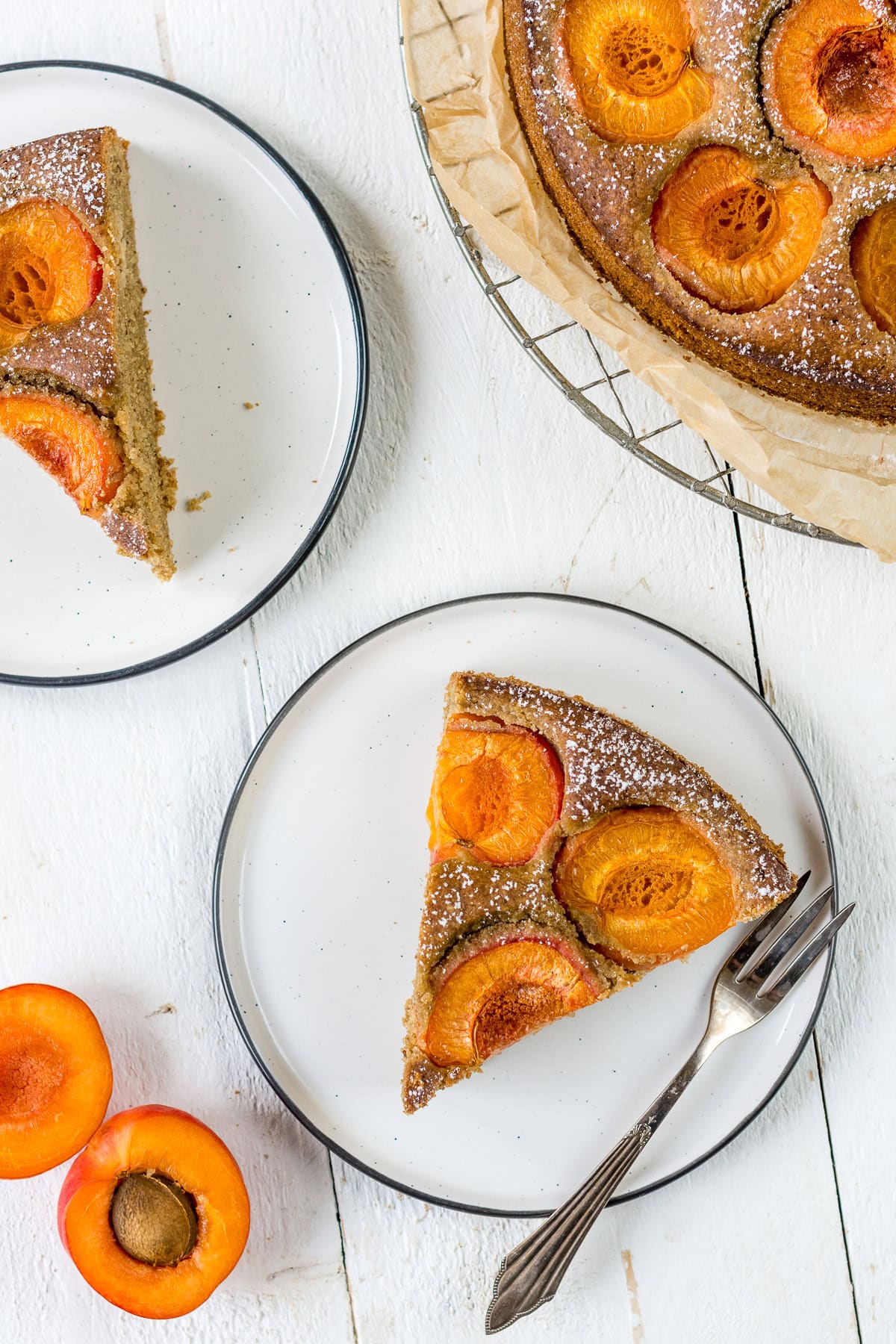 Zwei Stück Aprikosenkuchen auf weißen Tellern mit einer Gabel und frische Aprikosen.