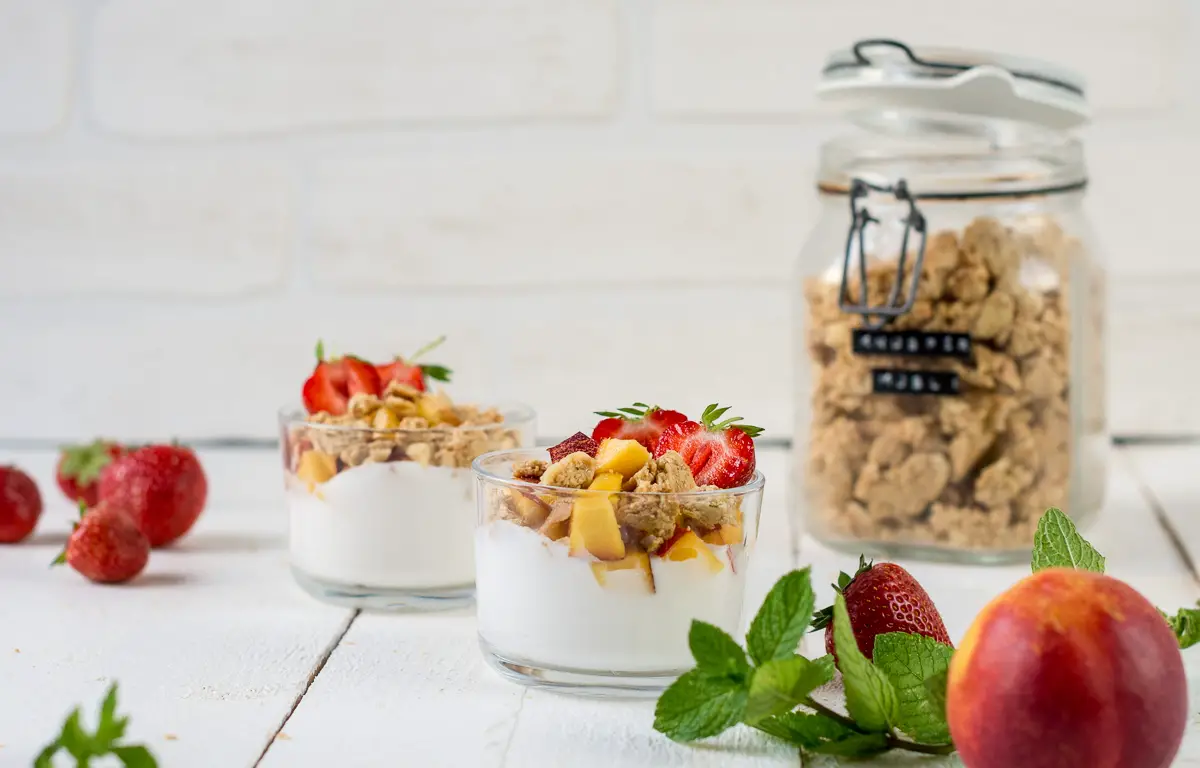 Zwei Gläser gefüllt mit Joghurt, frischen Früchten und Knuspermüsli. Im Hintergrund ein Bügelglas mit Müsli.