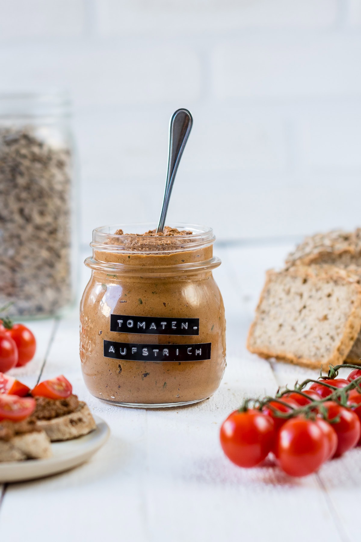 Glas mit Tomatenaufstrich und einem Löffel. Aufgeschnittenes Brot und frische Tomaten.