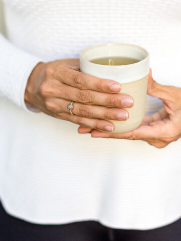 Person mit weißem Pullover hält eine Tasse Tee vor dem Bauch.