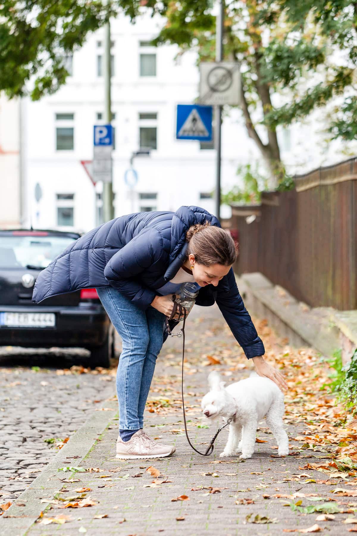 Frau beugt sich zu ihrem weißem Pudel und streichelt ihn auf der Straße.