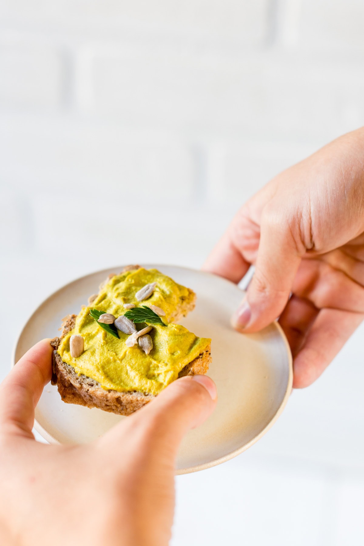 Pinne das Foto um dir das Rezept für veganen Curry Brotaufstrich zu speichern Person hält Teller mit Brot mit gelbem Curry Aufstrich, Sonnenblumenkernen und Petersilie