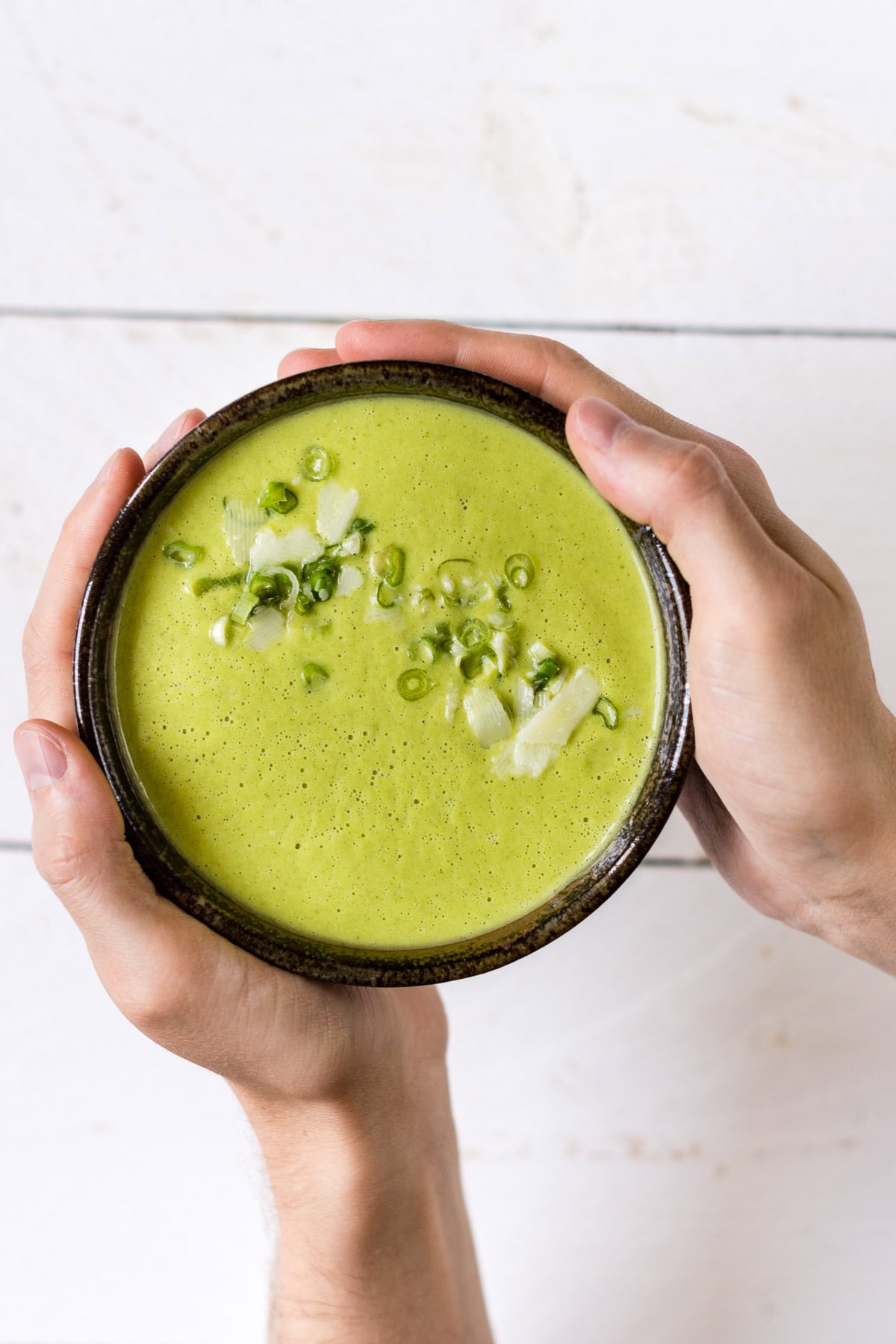 Pinne das Foto, um das Rezept für die Gemüsesuppe zu speichern! Hände halten eine Schüssel grüne Gemüsesuppe.