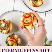 Hand hält einen Eiermuffin fest. Im Hintergrund sind zwei Teller mit weiteren Eiermuffins.