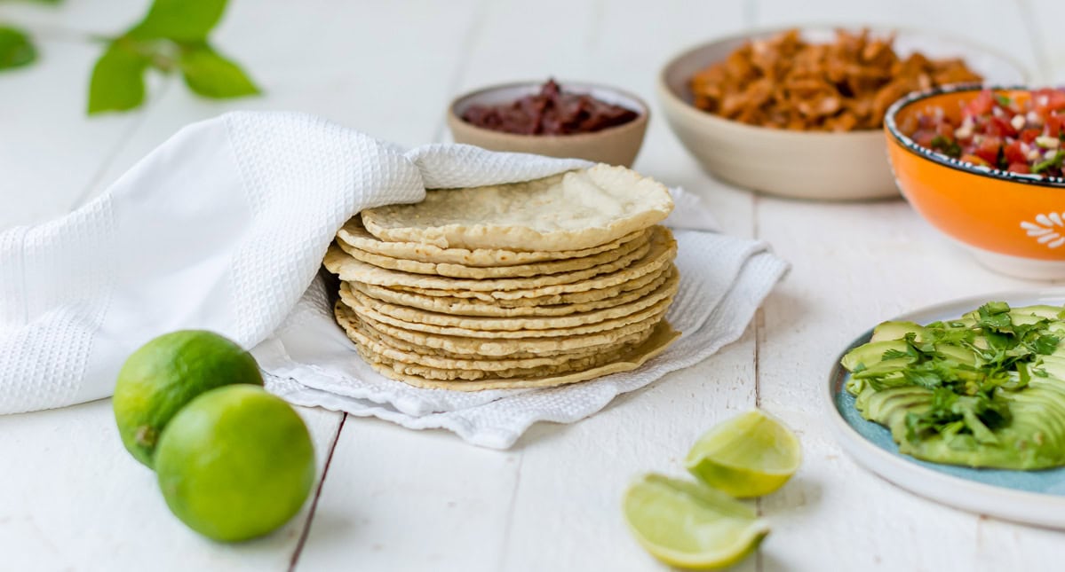 Frisch gebackene Tortillas in einem Tuch auf einem weißen Tisch umgeben von verschiedenen Zutaten zum Füllen der Tacos