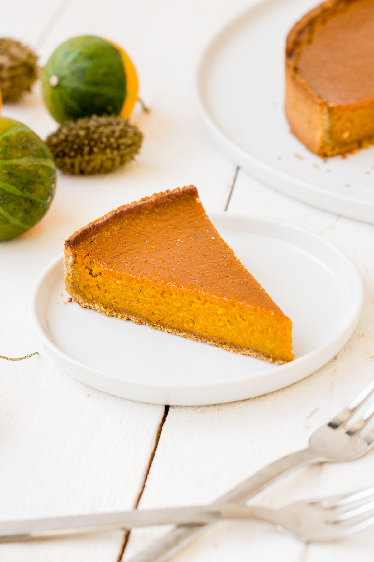 Foto von einem Stück glutenfreien Kürbis-Tarte (Pumpkin Pie) auf einem Teller mit Kürbissen und Kuchen im Hintergrund.