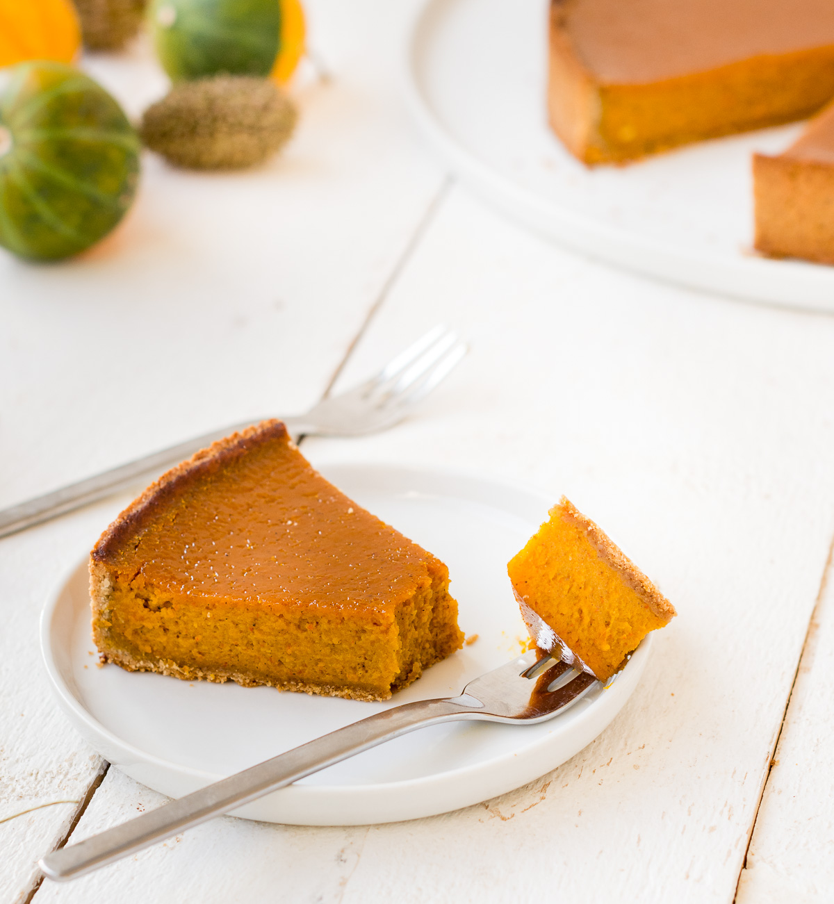 Foto von einem Stück glutenfreien Kürbis-Tarte (Pumpkin Pie) auf einem Teller mit Kürbissen und Kuchen im Hintergrund.
