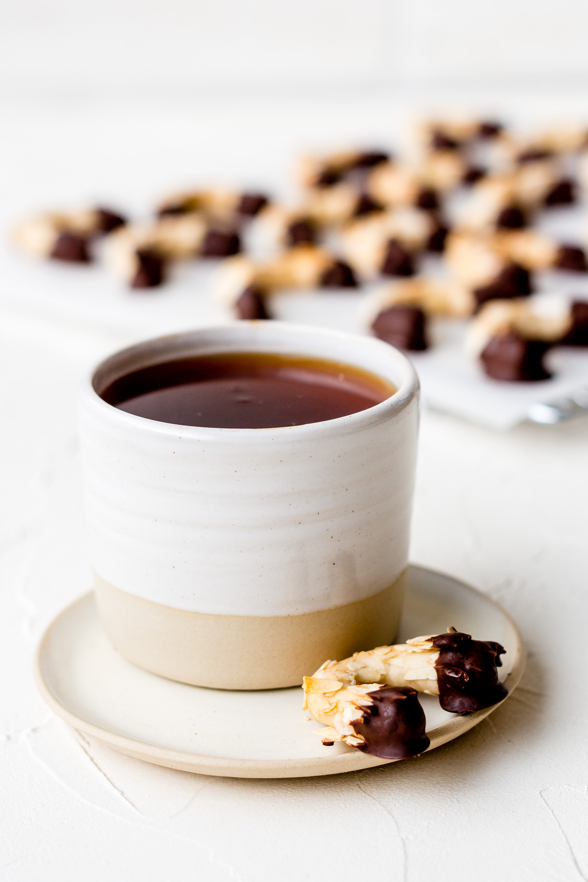 Tasse Tee mit MIni-Mandelhörnchen daneben und einem Blech Kekse im Hintergrund