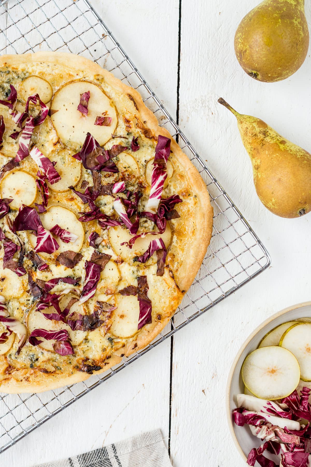 Topshot von einem Flammkuchen mit Birne, Radicchio und Gorgonzola auf Kühlrost, Birnen und Radicchio im Hintergrund.