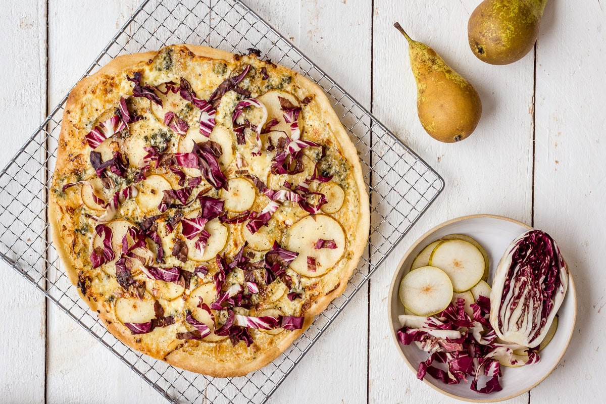 Topshot von einem Flammkuchen mit Birne, Radicchio und Gorgonzola auf Kühlrost, Birnen und Raddiccio im Hintergrund.