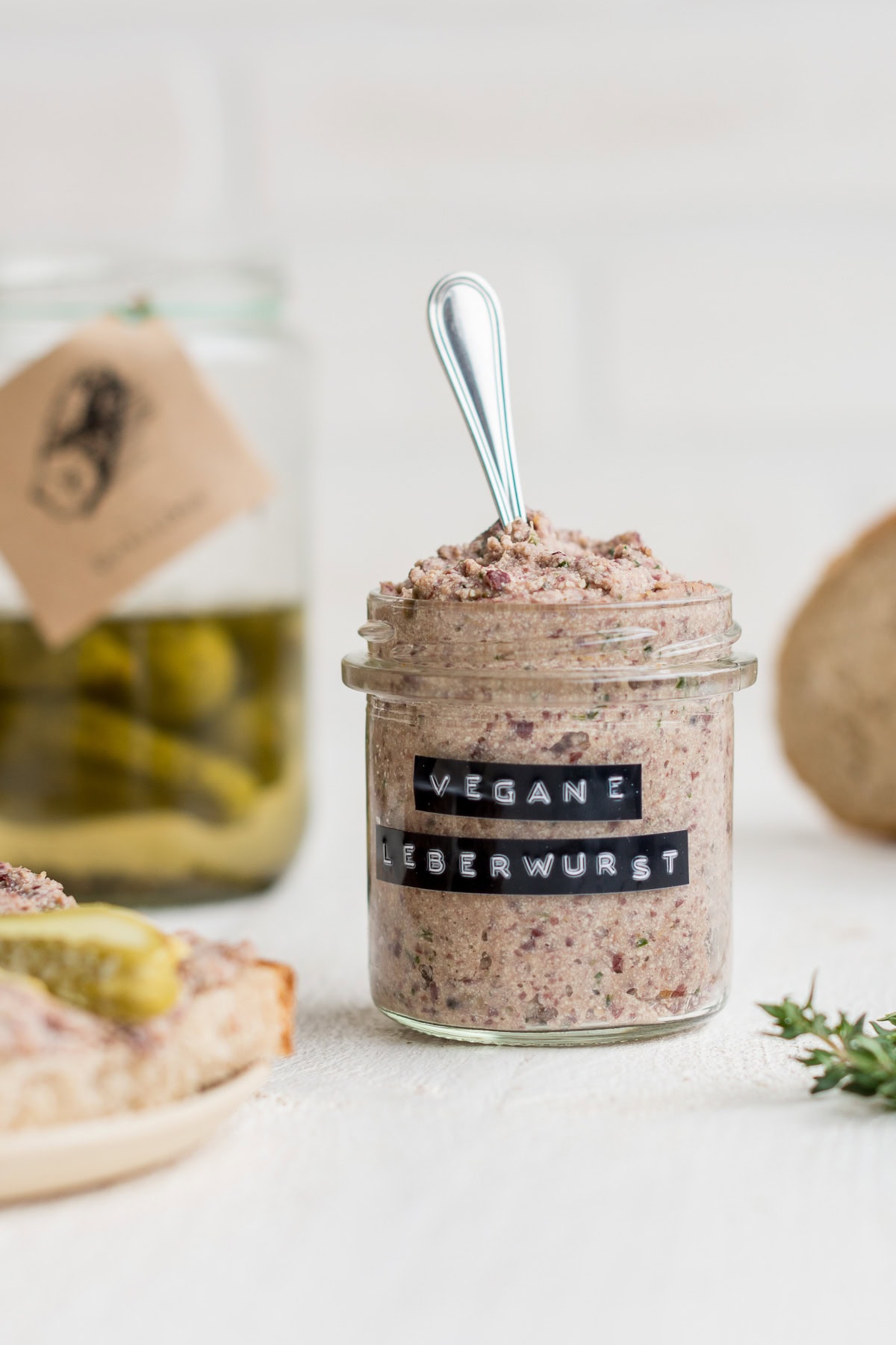 Ein kleines Glas mit veganem Leberwurst Aufstrich mit Gewürzgurken und Brot im Hintergrund.