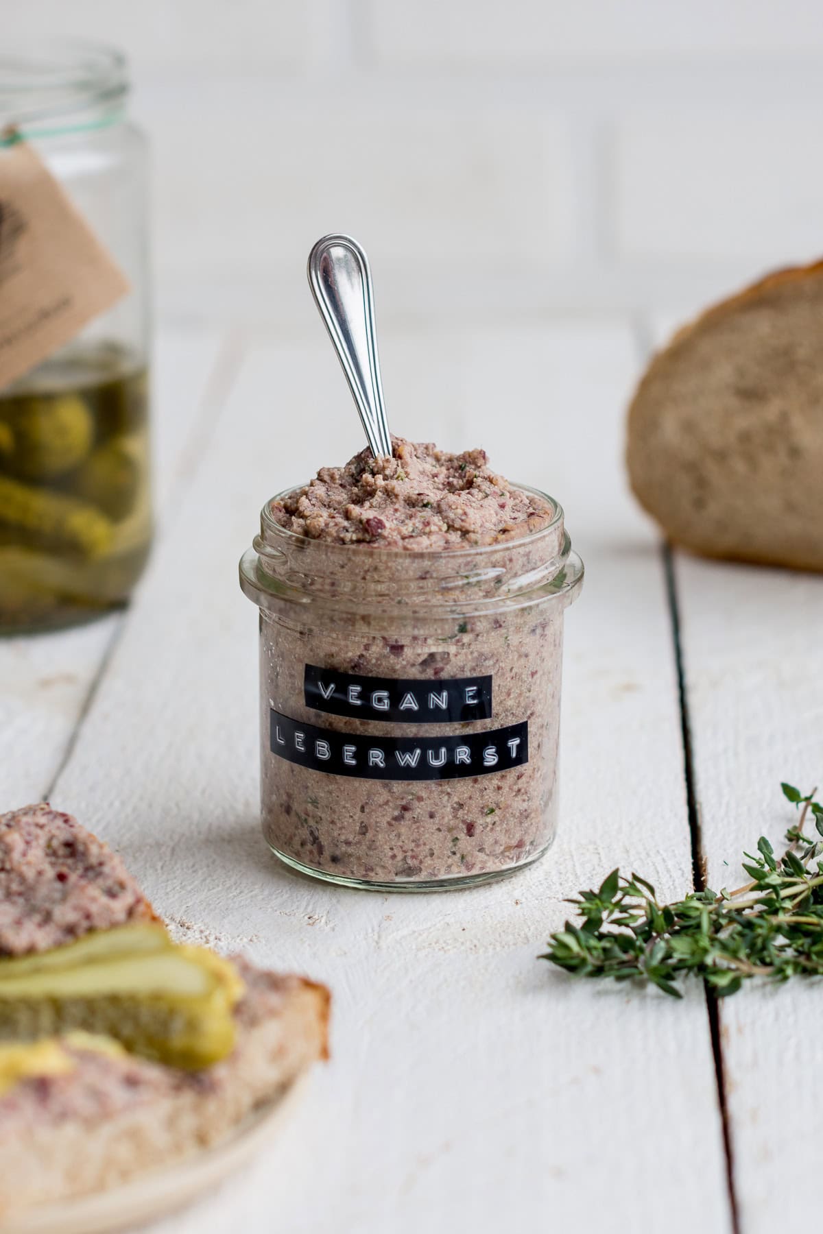 Ein kleines Glas mit veganem Leberwurst Aufstrich mit Gewürzgurken und Brot im Hintergrund.