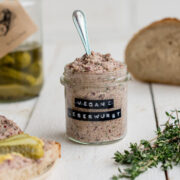 Ein kleines Glas mit veganem Leberwurst Aufstrich mit Gewürzgurken und Brot im Hintergrund.