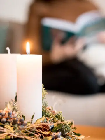 Adventskranz mit brennenden Kerzen im Vordergrund, im Hintergrund liest eine Frau eine Buch auf der Couch.